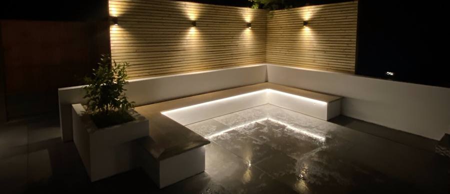 Garden terrace at night lit by recessed lights along a rendered wall