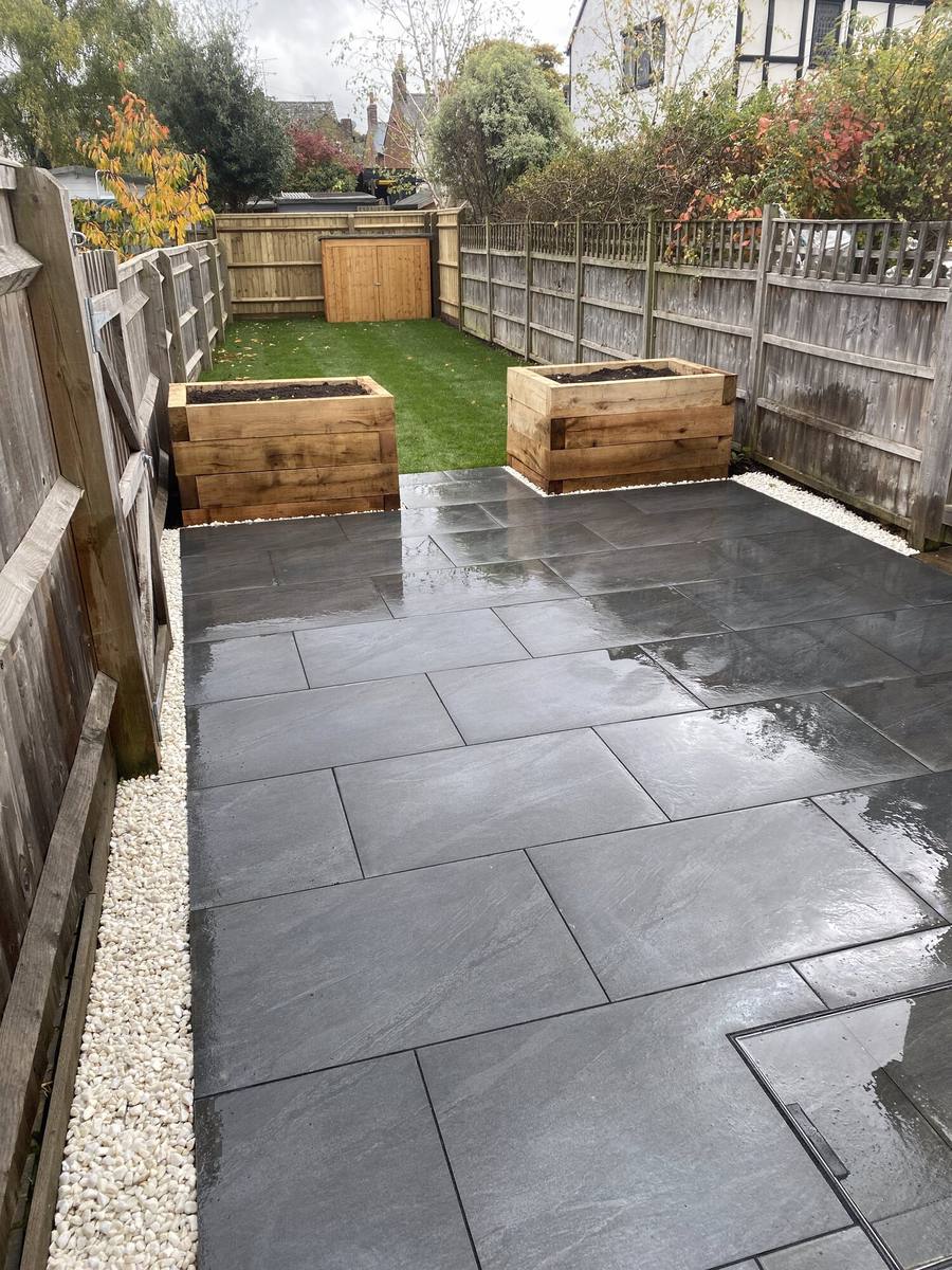 Grey porcelain patio in a narrow garden with two timber sleeper planters and artificial lawn beyond